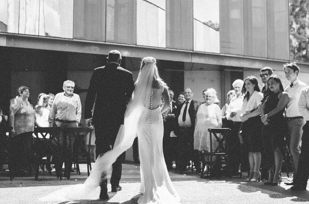 Black and white photo of a bride and groom in a paved outdoor setting with friends and family watching.