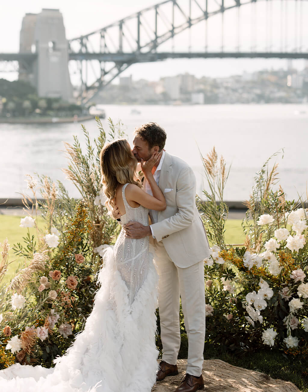 Bride and groom kiss on lawn with view of Sydney Harbour Bridge. Credit: Jimmy Raper