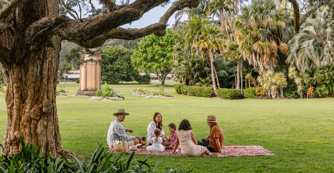 family on picnic rug on lush green lawns surrounded by shady trees